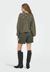 Maglione lavorato a maglia verde oliva con una consistenza spessa e motivi a diamante, abbinato a pantaloni corti in tweed grigi e stivali neri al ginocchio con fibbie.