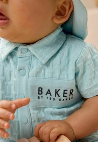 Camisa de manga corta azul claro con patrón geométrico texturizado, cierre de botones y "BAKER BY TED BAKER" impreso en negrita negra en la parte frontal.