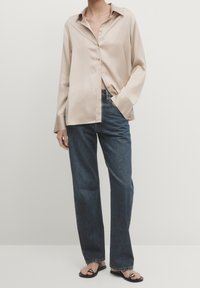 Personne portant une chemise en satin beige à manches longues, un jean droit bleu foncé et des sandales noires à bout ouvert avec un détail de chaîne argentée.