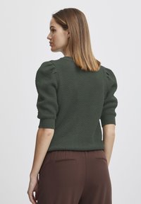 Groene gebreide trui met poffende mouwen en geribbelde manchetten. De textuur is verhoogd en patroonachtig. Gecombineerd met een paarse broek.