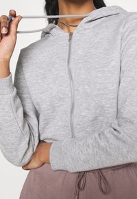 Sudadera gris con cremallera y textura suave, que cuenta con capucha, cordones y puños de canalé, combinada con pantalones suaves de color rosa claro.