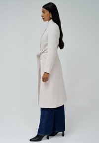 Cappotto lungo beige chiaro con cintura legata, tasche laterali e design slanciato, abbinato a jeans a gamba larga blu scuro e décolleté nere a punta.