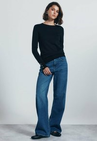 Maglione nero a maglia con maniche lunghe e rifiniture in bottone dorato, abbinato a jeans blu a vita alta e gamba larga, e scarpe slip-on nere.