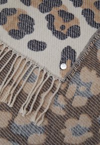 Écharpe douce en tissu tissé avec des motifs léopard beige, noir et marron, bords frangés, ornée d'une petite étiquette de marque ronde en métal.