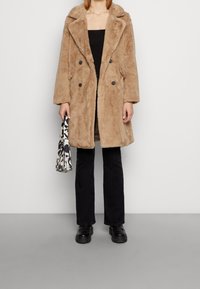 Beżowy płaszcz z sztucznego futra z dużymi klapami, zapinany na dwa rzędy guzik�ów, z czarnymi akcentami na guzikach, zestawiony z czarnymi spodniami i wzorzystą torbą.