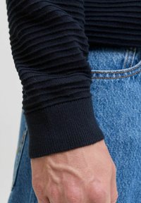 Manche de pull à côtes bleu marine foncé avec un design texturé. Le poignet est tricoté serré, associé à un denim bleu clair. Gros plan du vêtement.