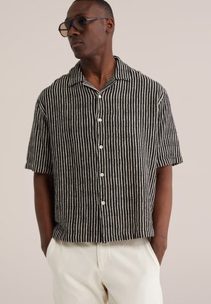 Mand iført sort og hvidt kortærmet skjorte med lodrette striber og knapper, hvide bukser og sorte briller, stående med hænderne i lommerne.