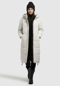 Polstret lang jakke i lys grå, med hette, frontglidelås, to sidelommer og svarte detaljer. Kombinert med en svart lue og topp.