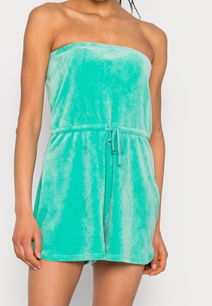 Turkis fløjl jumpsuit uden stropper med snøre i taljen, båret af en stående person, mod en ensartet lys baggrund.