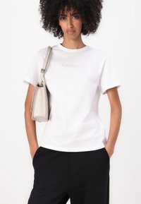 Witte katoenen t-shirt met korte mouwen en een ronde hals, voorzien van een subtiele grijze Calvin Klein-logo. Gepaard met zwarte broeken en een beige tas.
