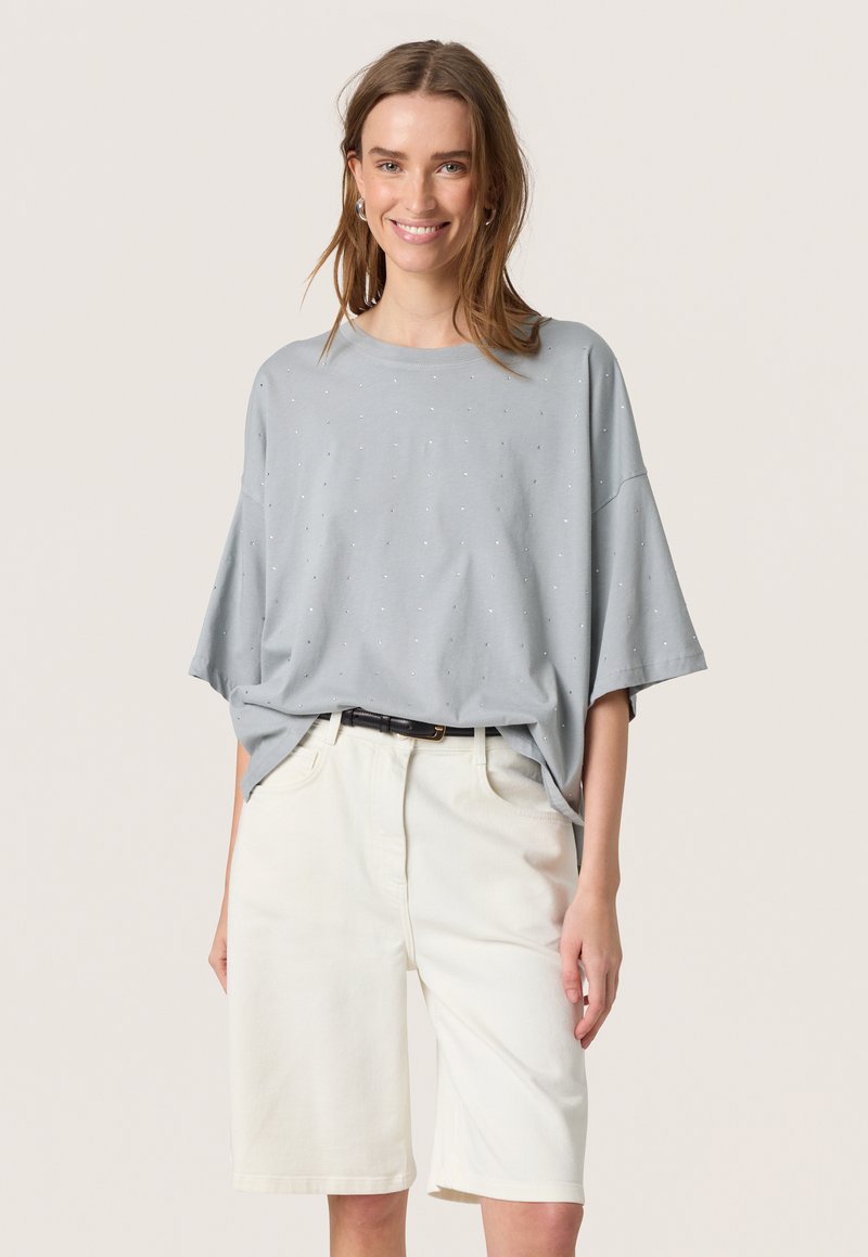 T-shirt oversize grigio chiaro a maniche corte, con piccoli punti decorativi, abbinato a pantaloni corti in denim bianchi fino al ginocchio e una cintura nera.