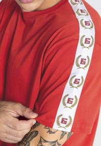 Detail fotografie osoby ťahajúcej rukáv červeného trička, ktoré má biely pruh s opakujúcimi sa zlatými vavrínovými listami a červeným logom "G".