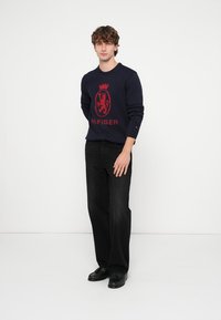 Maglione a maglia blu navy con logo rosso; abbinato a jeans neri a gamba larga e scarpe nere. Outfit semplice e casual con colori a contrasto.