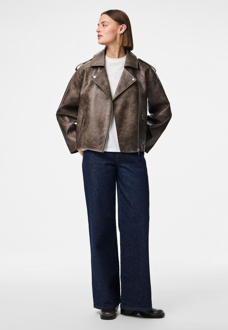 Giacca biker in ecopelle marrone con colletto a scollo e zip asimmetrica, abbinata a pantaloni di jeans ampi di colore scuro.