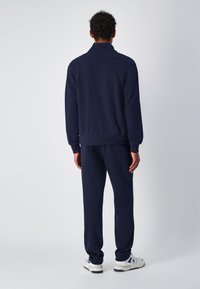 Tuta blu navy realizzata in tessuto morbido, con colletto alto e polsini a coste, indossato con scarpe da corsa bianche. Design semplice e confortevole.