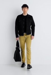 Νεαρός άνδρας που περπατάει, φοράει μαύρο σακάκι και παπούτσια, μουσταρδί παντελόνι, κρατώντας μια μαύρη τσάντα ώμου σε απλό λευκό φόντο.