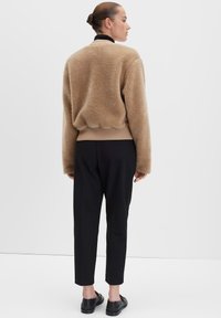 Pullover in pile beige accogliente con orlo a coste, abbinato a pantaloni neri. Tessuto morbido, vestibilità oversize e scollatura rotonda. Modello mostrato di spalle.