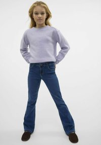 Jasnofioletowy sweter z długim rękawem wykonany z miękkiego materiału, zestawiony z niebieskimi dzwonami. Modelka stoi z rękami na biodrach, mając na sobie ciemne buty.