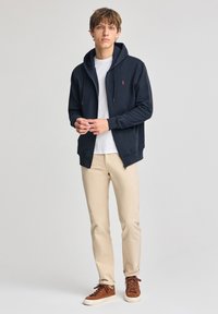 Sudadera con cremallera en color navy, con capucha ajustable, puños acanalados y un pequeño logo rojo en el pecho, combinada con pantalones beige y zapatillas marrones.