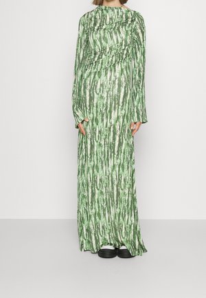 Vrouw die een lange groen-wit patroonjurk draagt met lange mouwen en zwarte platformschoenen op een effen grijze achtergrond.