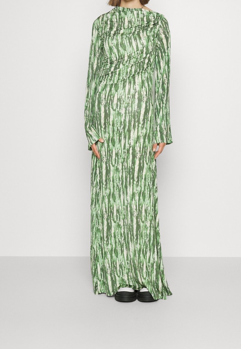 Femme portant une longue robe verte et blanche à motifs, à manches longues, avec des chaussures à plateforme noires sur un fond gris uni.