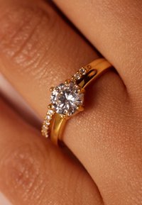 Goldring mit einem zentralen runden Diamanten, umgeben von kleineren Diamanten entlang des Bandes, vor einem strukturierten Hauthintergrund.