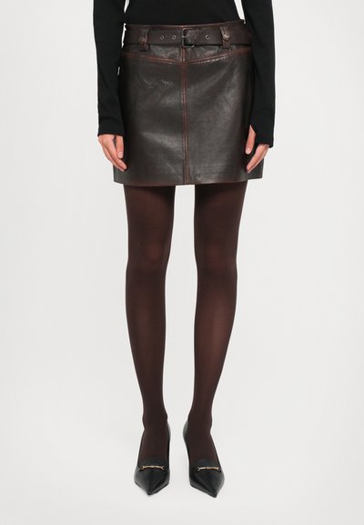 Vrouw draagt een donkergroene leren minirok met een bijpassende riem, zwarte doorzichtige panty's en zwarte puntige hakken met gouden accenten.