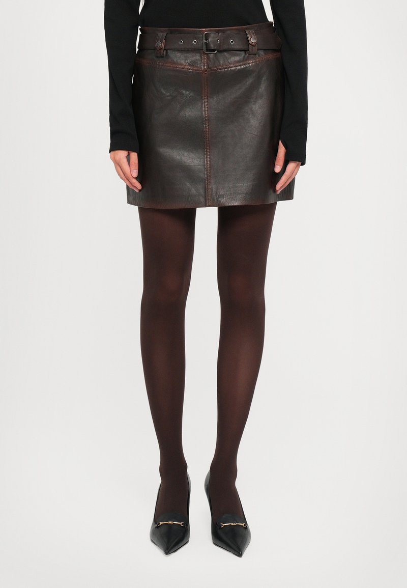 Woman wearing a dark brown leather mini skirt with a matching belt, black sheer tights, and black pointed-toe heels with gold accents.