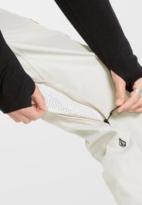 Pantalons de sport de couleur claire avec une fermeture éclair latérale révélant une doublure perforée, accentués de matériel noir et doré. Haut à manches longues noir.