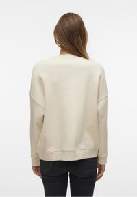 Sudadera color crema con un ajuste relajado, escote amplio y hombros caídos. Textura suave y dobladillo inferior acanalado, combinada con jeans oscuros.