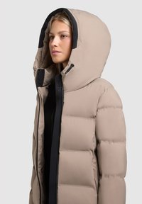 Beige pufferjack met een grote capuchon, een rits aan de voorkant en zwarte accenten. De jas heeft een gestikte textuur en een lichtgewicht ontwerp.