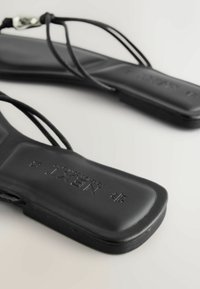 Sandales plates noires à bouts carrés et fines brides de cheville, taille 5 1/2, marquées de la marque "NEXT" sur la semelle intérieure, posées sur une surface blanche.