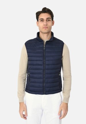 Uomo che indossa un gilet trapuntato blu navy con zip sopra una camicia beige a maniche lunghe e pantaloni bianchi, in piedi contro uno sfondo semplice.