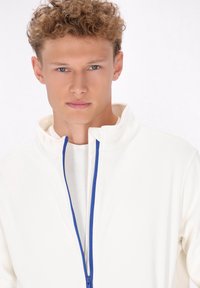 Giacca bianca con zip e colletto alto, dotata di zip blu. Il tessuto appare liscio con una consistenza leggera. Nessun motivo o accento notevole.