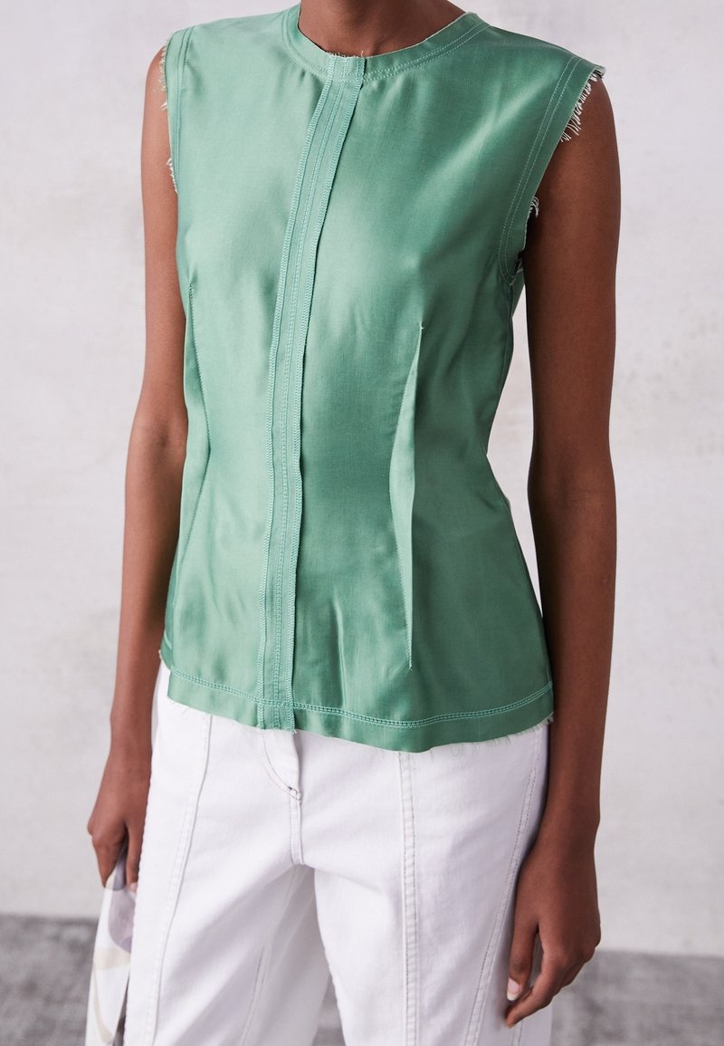 Haut vert sans manches avec plis sur le devant, porté avec un pantalon blanc taille haute sur une personne devant un fond neutre.