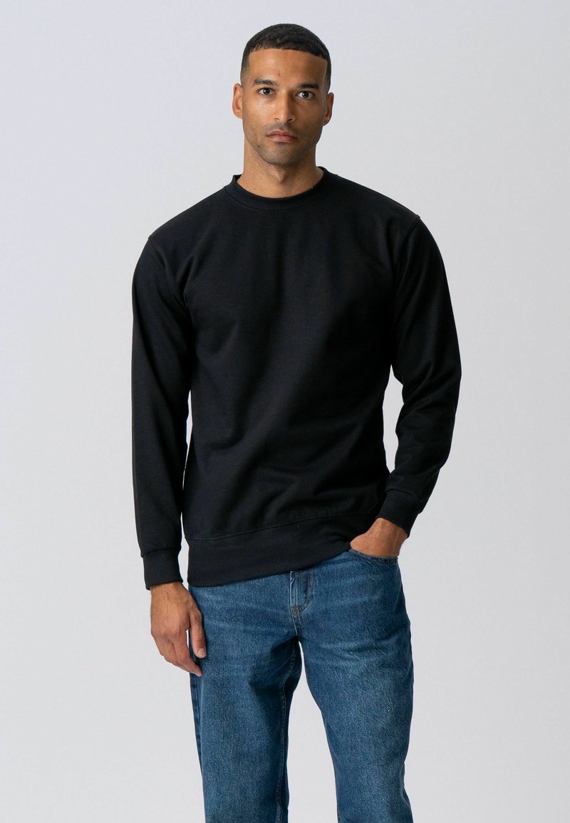 Homme aux cheveux courts portant un sweat-shirt noir à manches longues et un jean bleu, debout avec une main dans la poche devant un fond uni.