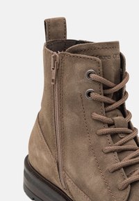 Brązowy zamszowy botek na kostkę z wiązaniem i bocznym zamkiem. Posiada teksturowaną powierzchnię oraz gumową podeszwę z delikatnymi beżowymi akcentami u podstawy.