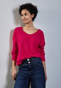 Maglione magenta a maniche lunghe con scollatura ampia, abbinato a jeans blu scuro con dettagli in bottone argentato e una vestibilità aderente con texture.