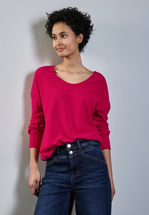 Maglione magenta a maniche lunghe con scollatura ampia, abbinato a jeans blu scuro con dettagli in bottone argentato e una vestibilità aderente con texture.