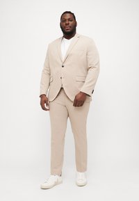 Beige tailored suit jacket and trousers with side pockets and two-button closure, worn with a white shirt and beige sneakers. Smooth texture.