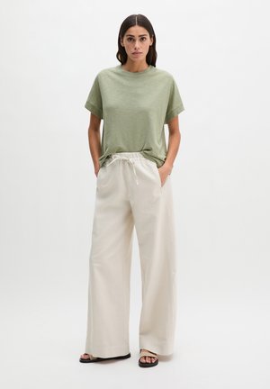 Femme portant un haut à manches courtes vert sauge, un pantalon beige à jambes larges avec cordon de serrage, et des sandales noires, debout devant un fond blanc uni.