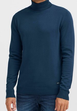 Man wearing a fitted navy blue turtleneck sweater and dark blue jeans, standing with arms relaxed by sides.