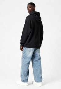 Felpa nera con cappuccio dal fit rilassato, abbinata a jeans baggy blu chiaro e sneaker bianche, mostrando un tessuto testurizzato e un design semplice.