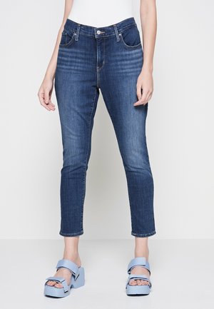 Kvinde iført mørkeblå skinny jeans og lyseblå platformsandaler står mod en ensfarvet baggrund.
