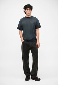 Zwart T-shirt met korte mouwen en een borstzak; gecombineerd met donkergroene brede broekspijpen en zwarte schoenen. Neutrale achtergrond, staande houding.