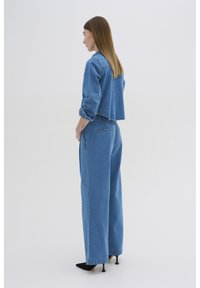 Conjunto de mezclilla que incluye una chaqueta azul corta con dobladillo deshilachado y unos pantalones vaqueros de pierna ancha a juego. Unos zapatos de tacón negro completan el look.