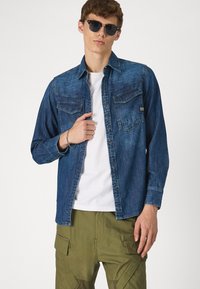 Tumši zilas džinsu krekls ar apkakles kaklu, divām krūšu kabatām un pogu aizdari. Saskaņots ar olīvu zaļām kravu biksēm.