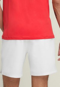 Rotes Sportshirt mit glatter Textur, kombiniert mit weißen Shorts, die ein einfaches Design und einen taillierten Schnitt haben. Sichtbare Nähte am Saum.