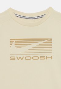 Kreminės spalvos medvilninis marškinėliai su tekstūruotu auksiniu logotipu ir horizontaliomis linijomis virš žodžio "SWOOSH" ryškiu, aiškiu šriftu.