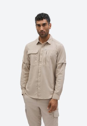 Muškarac odjeven u bež košulju dugih rukava s gumbima i džepovima na prsima te odgovarajuće bež cargo hlače, stoji nasuprot jednostavnoj pozadini.